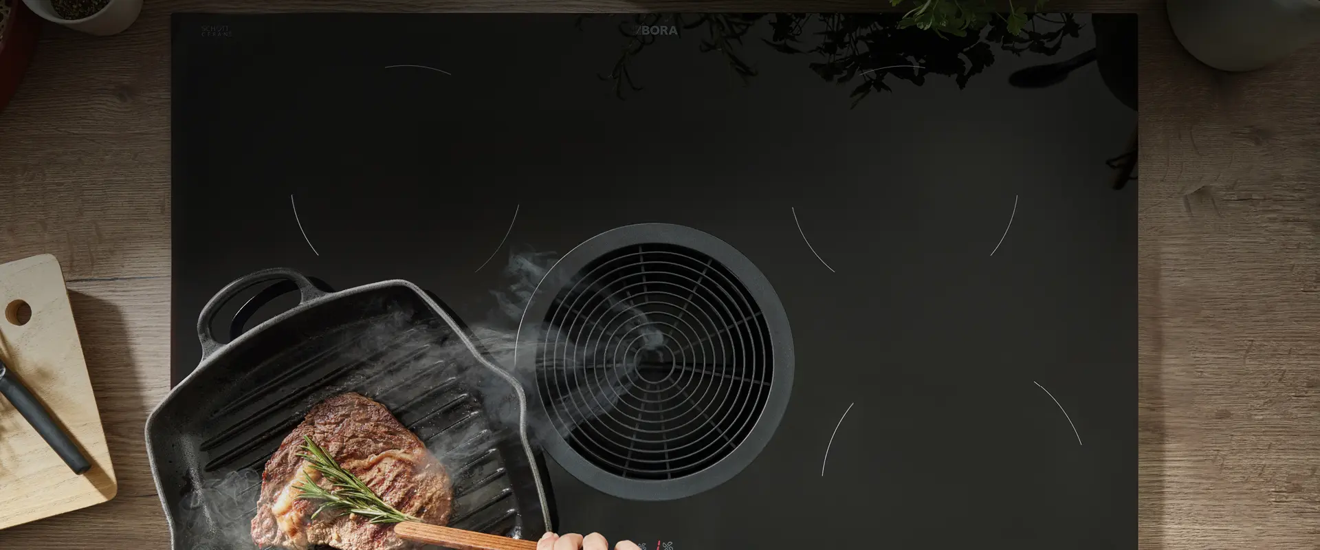 Un steak saisit sur une poêle à griller en fonte noire, à côté du ventilateur d'extraction central d'une élégante plaque à induction noire. Un steak saisit sur une poêle à griller en fonte noire, à côté du ventilateur d'extraction central d'une élégante plaque à induction noire.