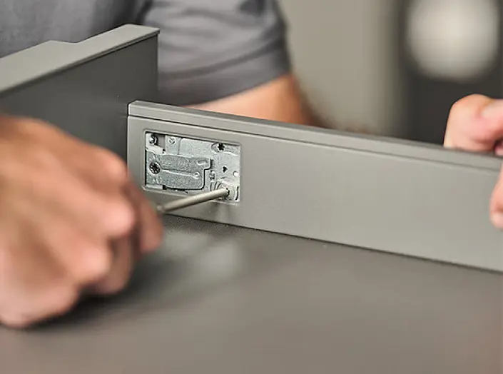 Une personne resserre une charnière métallique sur un tiroir gris à l'aide d'un tournevis. Une personne resserre une charnière métallique sur un tiroir gris à l'aide d'un tournevis.